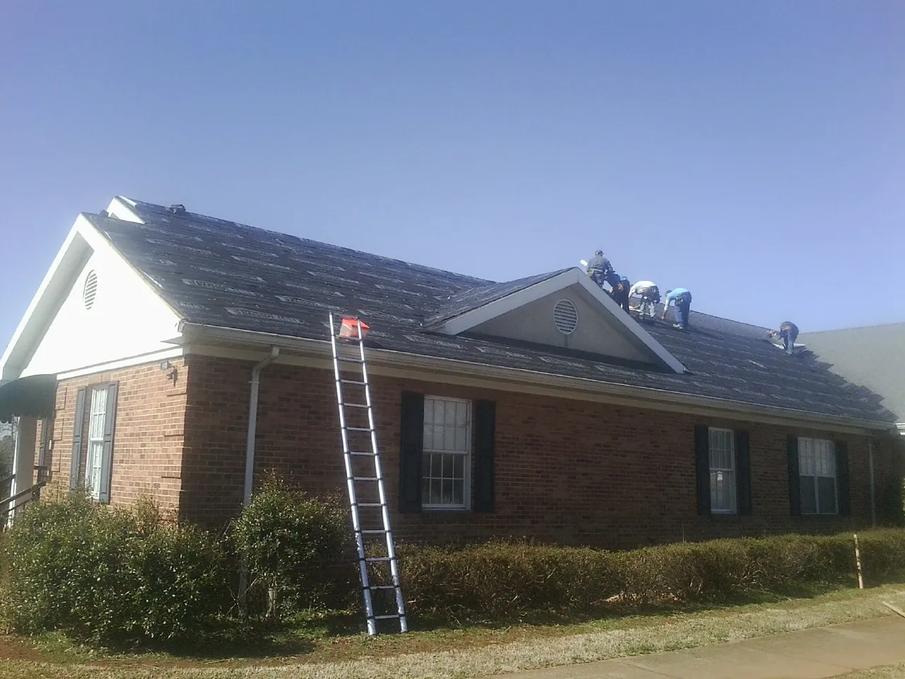 Crew tearing shingles off a brick ranch roof with a ladder set — Metal Roof Repair work by our crew in Halstead