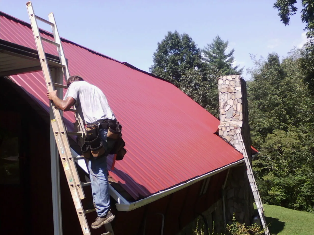 Roofer on a ladder beside a red metal roof and stone chimney — Soffit & Fascia Repair by Roofing Brothers in Halstead