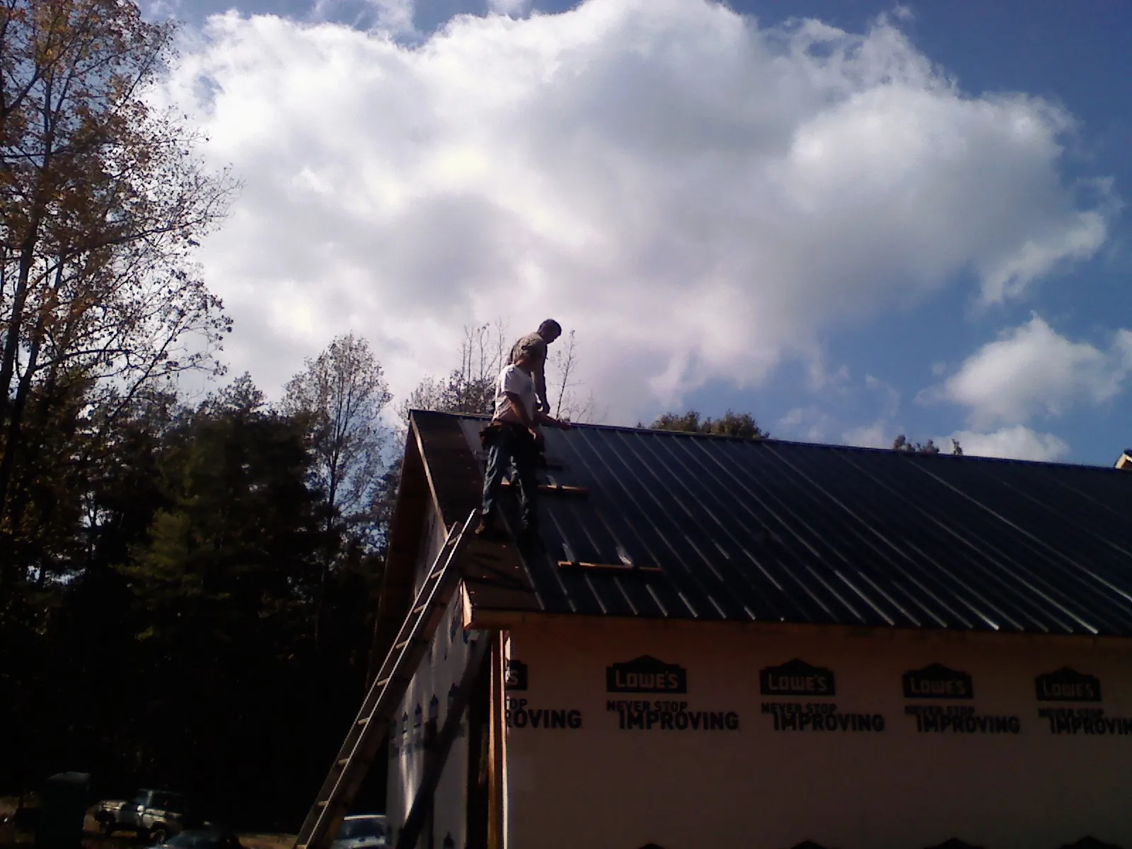 Roofer fastening dark metal panels high against a cloudy sky, from a Roof Inspection job in Halstead